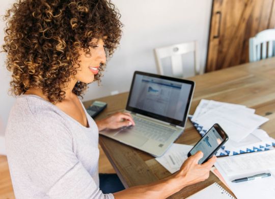 A woman sits at her dining room table with laptop and financial reports doing her monthly budget. She is smiling at the ease of use as she works on her smart phone banking app to do monthly finances, pay taxes and save money for the future.